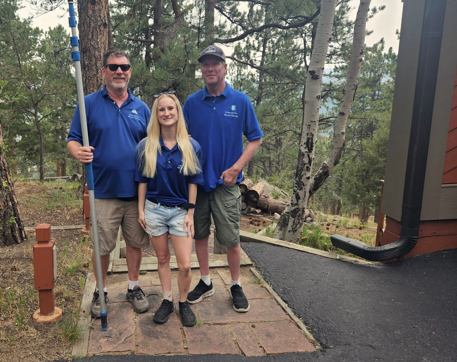 Three of the crew in branded polo shirts standing on a flagstone path outside a home in the pines, one holding a water-fed pole.
