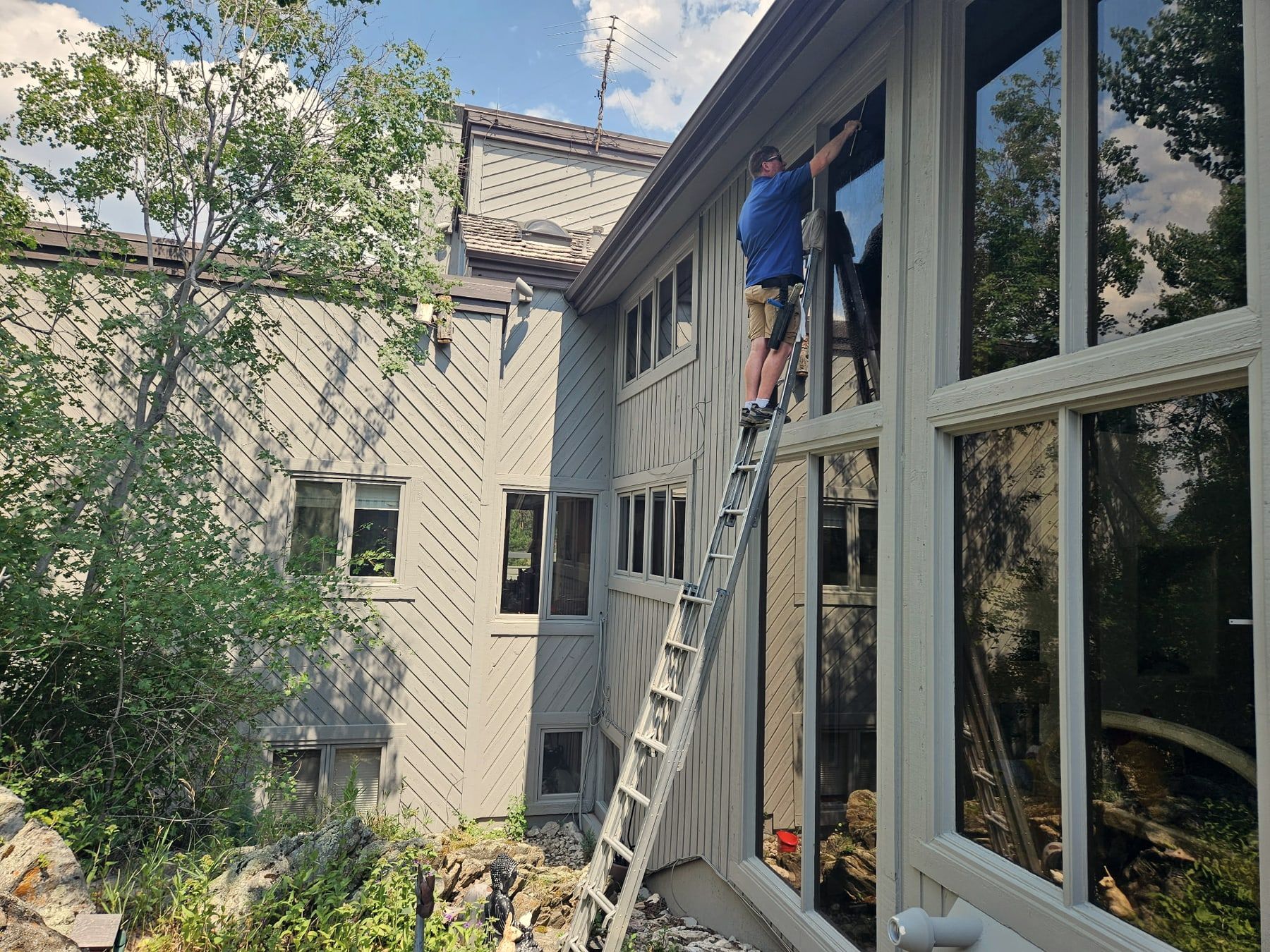 One of the crew on a ladder cleaning the upper panes of a two-storey wall of glass, seen along the side of the house.